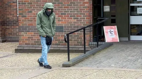 Christopher McEwen walks near the entrance of a building. He is wearing light blue jeans, with a green jacket with the hood up. He has a scarf covering the lower part of his face. He is hunched over with his hands in his pockets. 