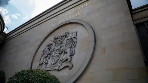 Exterior view of the High Court of Justiciary in Glasgow, showing the words The High Court of Justiciary carved into the sandstone façade above a large stone relief of the Royal Coat of Arms. Photographed from a low angle, the court building fills the frame, with greenery in the foreground and a partly cloudy sky overhead.