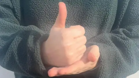 BBC A close-up of a person signing - it shows a flat palm, with the other hand placed on top giving a thumbs up, the person is wearing a grey fleece top.