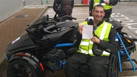 A man in a wheelchair wearing a hi-vis jacket. He is in front of a black motor trike and holding a piece of paper.