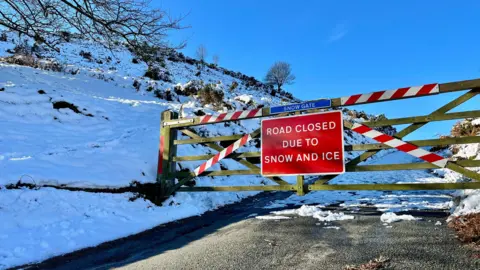 BBC Weather Watchers/Snapper Simon A five-bar wooden gate across a road with a road sign "Road closed due to snow and ice". A snowy landscape lies behind, with compressed snow on the carriageway. 