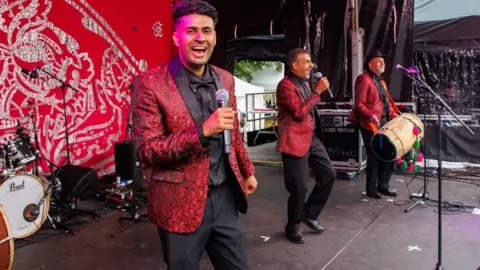 Supplied The Masti Band left to right: Aamir Rana, 36, holding a microphone, his father Amjad Rana, 66, also holding a microphone and his uncle Surinder Sandhu, 62, with a dhol drum - all wearing decorative suits and performing on stage in Glasgow in July 2026.