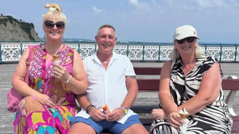 Two woman and a man sat on a bench on Penarth seafront. They are each holding an ice cream. The two women are wearing summer dresses with short sleeves. The man sat between them is wearing a short-sleeved button down shirt and shorts.
