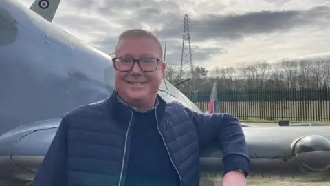 Andrew Pawson stands in front of a WW2 style fighter plane. He has short brown hair, brown glasses, and wears a blue puffer jacket and a blue jumper.