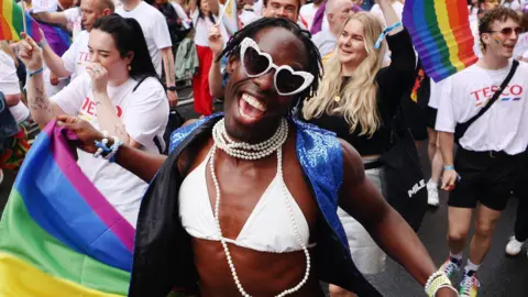 A smiling woman in a cream bikini top, wearing heart-shaped sunglasses and a string of pearls is in the middle of a parade of people. She has an LGBT flag in her hand. 