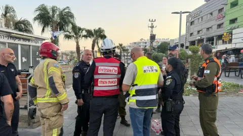 Israel Defense Forces Emergency services at the site of a drone strike in Eilat, Israel. They are standing in a circle with palm trees and some buildings behind them.