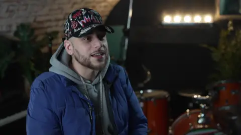 JT, a man in his thirties wearing a blue puffer coat and a grey hoodie. He is wearing a black hat with graffiti printed on it. He has a bit of facial hair and is smiling. The background is blurred but a drum kit can be seen, along with lights and a large green cheese plant.