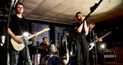 All band members are dressed in black. Luke, the guitarist is holding his white and black electric guitar. Joe, the drummer, is in the backdrop holding his sticks. Front man Ben is grasping the microphone, whilst bassist Ed is clutching his bass guitar.  