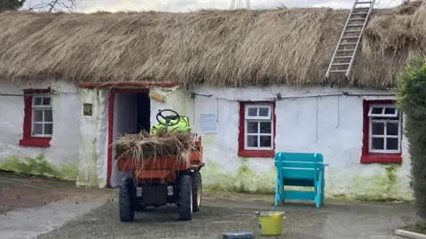 Doagh Famine Village A thatched cottage with a red door and a window either side of the door. The cottage is painted white. a small dumper truck sits in front of it