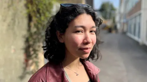 A woman with shoulder-length dark hair smiles at the camera. She wears a dark red jacket and has sunglasses propped up on her head.