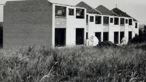 GaelStair project A black and white image of a row of terraced houses being built, and there is long grass in the foreground.