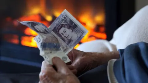 A close-up image showing an elderly person wearing a blue jumper holding two £20 notes out on front of them. In the background there is a burning fire out of focus.