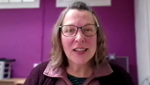 A woman with shoulder-length, brown hair looks towards the camera. She is wearing glasses, a pink shirt and a purple cardigan. She is sitting in front of a purple-coloured wall.