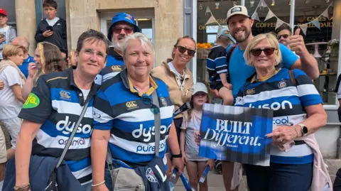 Seven people, five of whom are in Bath shirts for the parade. They are standing in front of a coffee shop with coloured bunting in the window