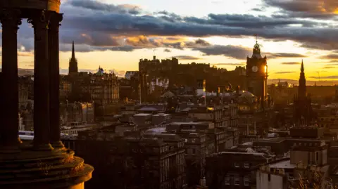 STEVEN NEISH A general view of Edinburgh's New Town from Calton Hill. A monument is on the left of the picture. The new town can be seen below at sunset. The sky is orange with streaks of blue-grey cloud.