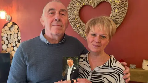 A man and a woman looking at the camera. The man, on the left, is wearing a blue jumper. He has his arm round the woman, who has short blonde hair and is wearing a zebra-print shirt. They're standing in front of a wall which is painted red. To their left are a number of logs which sit in the wall, and behind their head is a wicker heart decoration. They are holding a small photo of a man wearing a wedding suit. 
