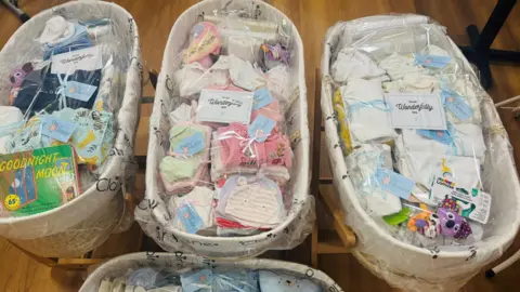 Baskets of baby clothes and toys. There are wrapped to look like hampers. The floor is made of wood. 
