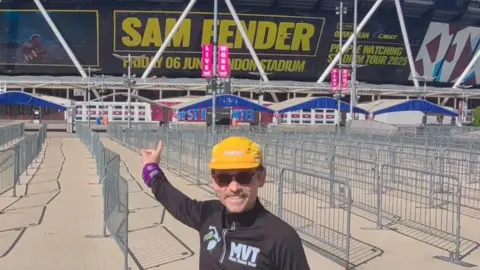 Andy Hobson, dressed in a running top and a yellow hat, is pictured standing outside the London Stadium. The stadium side reads "Sam Fender" in large yellow letters, as well as carrying the date of his performance. 