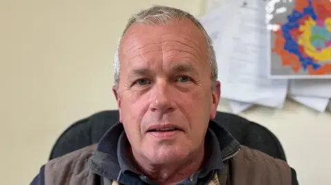 Matthew Cowan has short grey hair. He is sitting in a chair at his desk, wearing a brown gilet over a navy jumper.