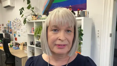 Kerry has a short gray haircut, an eyebrow piercing and blue eyeliner. She looks directly into the camera and stands in front of a cupboard with plants and toys on it. 