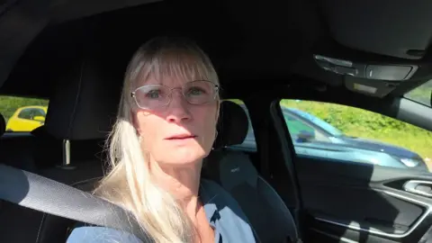A blonde woman wearing glasses sits in a car frustrated by the delays caused by the rescheduled roadworks.