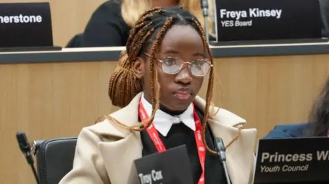 A black teenager with long black cornrows in her hair, dyed brown for much of their length, wears a cream coat and a black jumper, with a red lanyard. She is sat at a desk with a microphone in front of her, to one side