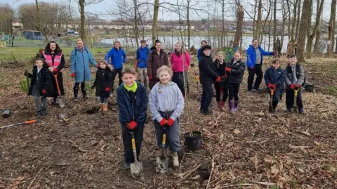 North Tyneside Council School children in Killingworth Lakeside Park during a visit to plant trees. A boy and girl are standing at the front of the group and are each holding a spade. A potted tree stands next to them. A group of children and teachers stands behind the. In the background are existing trees, a lake and a play park.