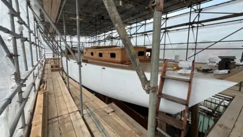 A white ship under construction surrounded by scaffolding and a ladder. 