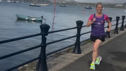 Jo wearing her purple Sands T-shirt and black shorts. She is running beside the River Tamar with black railings to her right. There are boats on the water beyond the railings.
