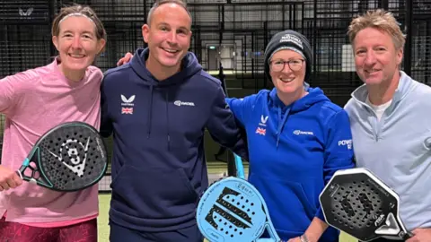 Two sportswomen and two sportsmen are standing on a padel court in a midshot, smiling to camera and each holding a padel racket