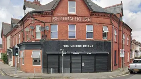Google A shop front on a red brick building is painted black with white writing which reads: The Cheese Cellar