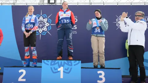 Special Olympics Three women in skiing gear on a blue podium with 2, 1, and 3 in white numbers below where they are standing. Each woman has a medal around her neck and is smiling. A grey-haired woman in a white jacket, sunglasses and black trousers to the right of the podium is clapping.