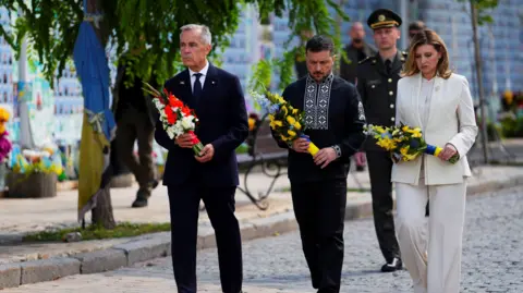 The Canadian press is carried through the Prime Minister of AP Canada Mark Carne, and his short gray hair and face, red and white flowers walking in a dark suit on a street in Kiev, Ukraine. Ukrainian President Voludmir Zelinsky, his hair and beard is dark and short, is wearing black Ukrainian clothes with white embroidery around the collars and in the lower line. His wife, Olina Zelinska, wears a white suit. The couple wears bleak expressions and both carry yellow and blue flowers. A soldier walking in the background. 