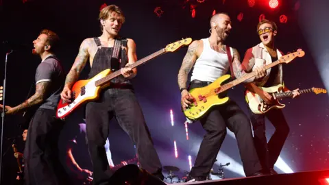 Getty Images L-R- Danny, Dougie, Matt, Tom Dougie, Matt and Tom are playing electric guitars. Danny is standing in front of a mic singing, wearing blue trousers and blue striped shirt. Dougie wears black dungarees. Matt is wearing black jeans and a white vest. Rom wears all black with a white jacket. 
