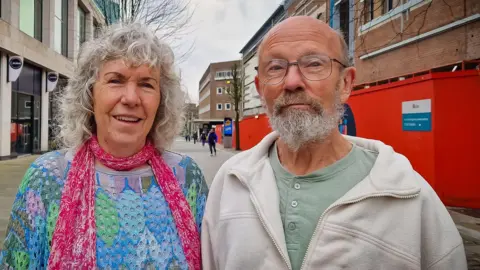 The Wallich Valerie and Chris standing together on a street in Swansea. She is wearing a brightly coloured crochet top and pink scarf. He is wearing a beige fleece jumper and green button down shirt. 