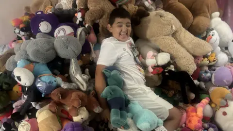 Send Community Connected Jenny's son Junior surrounded by lots of soft toys. He smiles widely.