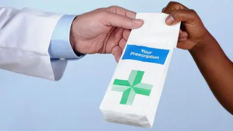 A stock close image of a pharmacists hand passing over a white paper bag with a green cross on it. They are wearing a white coat and blue shirt, another arm grips the bag receiving it