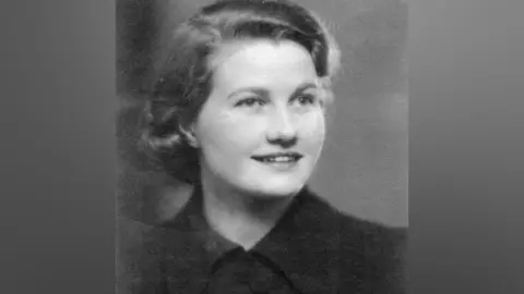British Resistance Museum A black and white studio-style photograph of Thea Ward, a young woman with fairly short hair, turning slightly to look to the right and smiling. She is wearing a dark-coloured shirt done up to the neck. 
