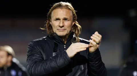 Gillingham head coach Gareth Ainsworth head shot clapping fans in winter waterproof blue coat