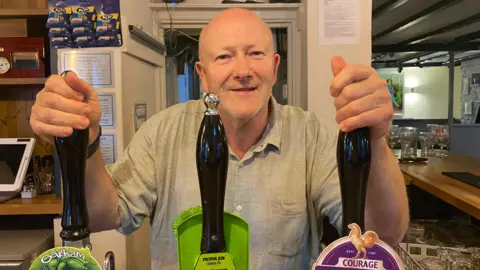 BBC Keith Marsden wearing a grey t-shirt and holding the pumps of beer