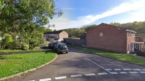 A view of Ashfield Close, Exmouth. There is a large tree to the left of the entrance with two cars parked at the side of the road. The houses are terraced. 
