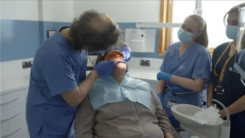 A dental patient lies in a chair as a dentist works on her teeth. Two other dental staff who all wear masks watch on.