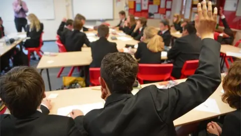 BBC The backs of teenagers in a classroom with a boy raising his right hand