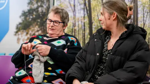 Cambridge University Hospitals Shirley, a woman with greyish hair, wearing a black jumper with colourful motifs and blue-framed glasses, sitting on a purple chair, knitting, while her daughter in a black coat, is sitting next to her watching Shirley.