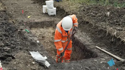 East West Rail A person wearing high-visibility orange protective clothing and a white safety helmet is working inside a carefully marked excavation trench. The rectangular pit is outlined with string lines and surrounded by mounds of excavated soil. Tools, containers, and sample bags are positioned around the site, suggesting a structured digging or surveying process. The scene appears to show an archaeological or construction-related excavation being carried out in an outdoor area.
