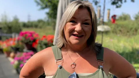 Jennie Franklin standing in her garden with bright flowers seen behind her. She has fair hair to her shoulders and is wearing a green dungaree-style top.