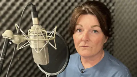 BBC Gayanne is in a recording booth with black foam panels. She is sat beside her silver microphone which his hanging upside down with a black pop shield in front. She has brown hair, blue eyes and is wearing a blue top that says "The Bubbling Toad."