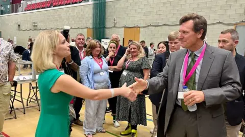 Labour's Charlotte Holloway on the left in a green sleeveless dress is shaking hands with Angus Forbes on the right who is wearing a grey suit and an olive tie with a pink lanyard and holding a bottle of water. Campaigners are in the background in the sports hall of the Plymouth Life Centre.