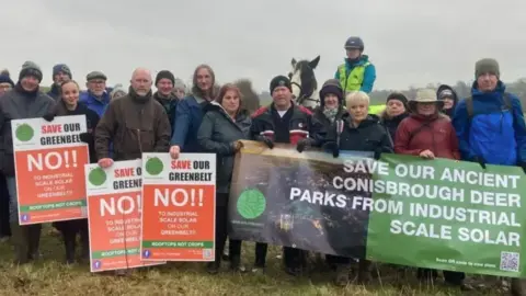 Several people pose for a photo behind placards and banners protesting against the solar plant plans. One reads 'save our green belt'. They stand in a field.
