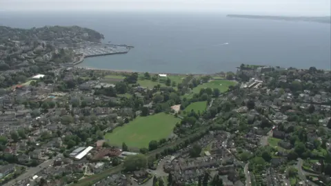 A drone shot from high in the sky overlooking Torquay, Devon. There is lots of buildings and green spaces as well as the sea in the distance.